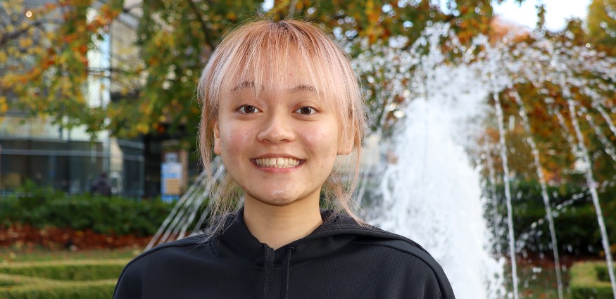 Female Asian student wearing a black hoody standing in front of a water fountain.